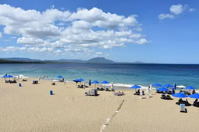 a beach with blue umbrellas and chairs and the ocean at Alicia Beach, Hispaniola Sol! in Sosúa