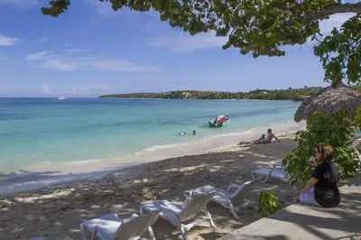 a woman sitting on a beach with chairs and a boat in the water at Paradise Island Beach Resort in Punta Rucia