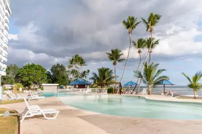 a swimming pool with palm trees and the ocean at Luxury Beachfront 2BR at Las Olas, Juan Dolio in La Puntica de Juan Dolio
