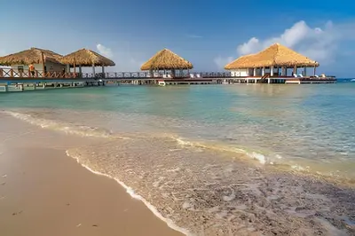 a beach with two thatch huts and the ocean at Club Hemingway Amplio Apartamento en la Playa de Juan Dolio in Los Corrales