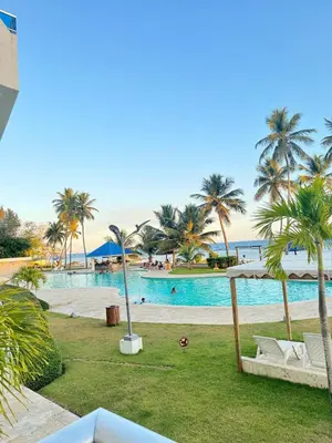 a swimming pool with palm trees and a beach at Las Olas Apartment, Juan Dolio in Guayacanes